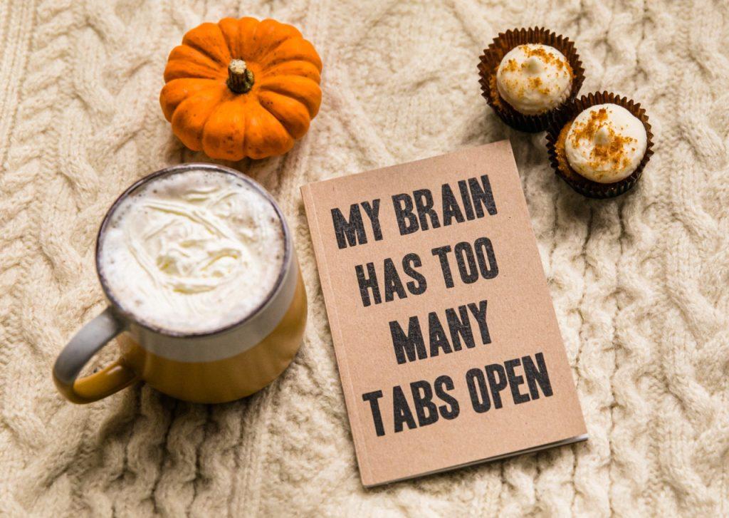 Auf hellem Strick liegt ein Kürbis, ein Muffin, ein Kaffee und ein Notizbuch mit dem Titel "My brain has too many tabs open"